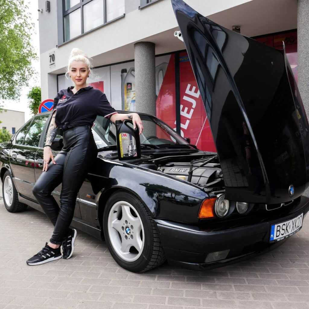 Dziewczyna stoi przy klasycznym BMW i prezentuje olej silnikowy Millers 5w40. Rozważa wybór 5w30 czy 5w40