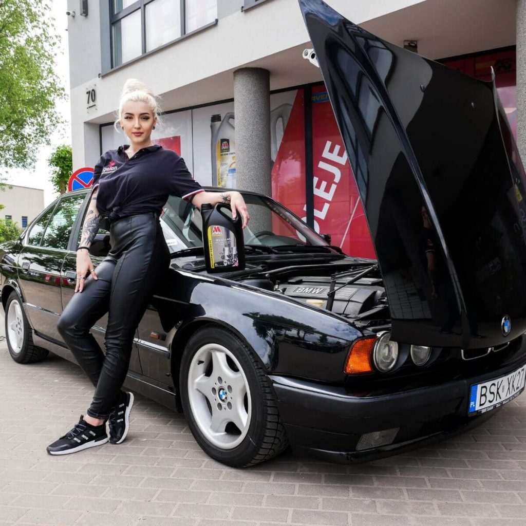 dziewczyna o blond włosach stoi oparta o auto marki BMW i trzyma olej Millers. Millers oil to marka, która również ma w swojej ofercie oleje PAO. 